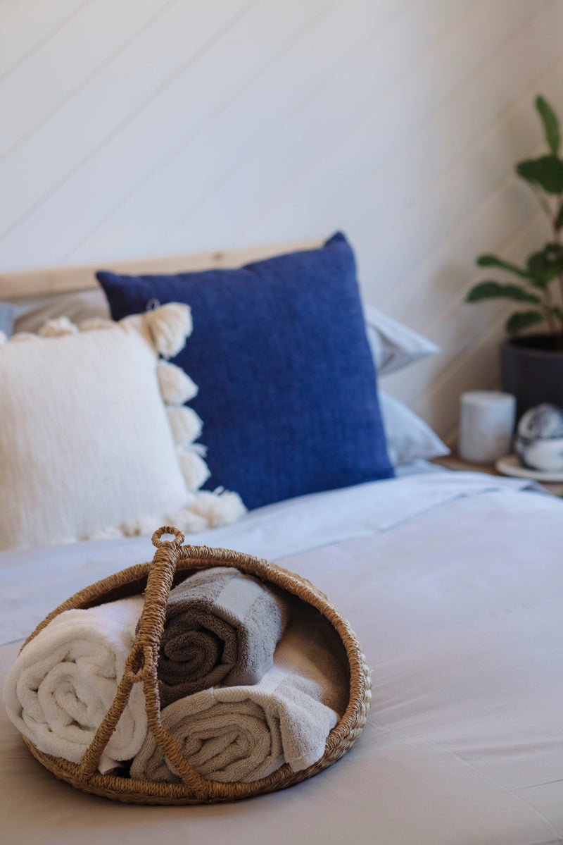 Basket with folded blankets on a bed with decorative pillows and a plant in the background